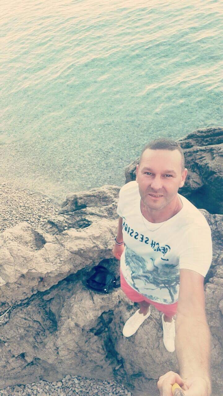 Mężczyzna robiący selfie na skalistym wybrzeżu z widokiem na turkusowe morze, ubrany w białą koszulkę i czerwone spodenki.