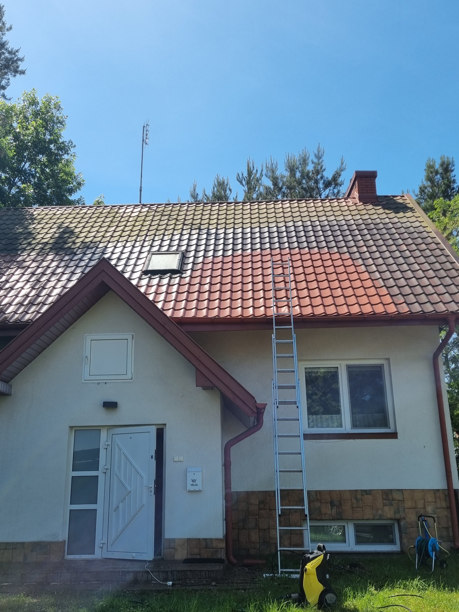 Częściowo umyty, czerwony dach domu z drabiny. Widoczny sprzęt do mycia ciśnieniowego i fragment trawnika. Błękitne niebo w tle.