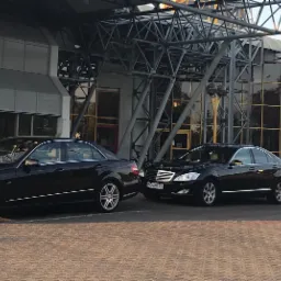 Trzy czarne limuzyny Mercedes-Benz stojące w rzędzie przed nowoczesnym budynkiem z metalową konstrukcją i szklaną fasadą.