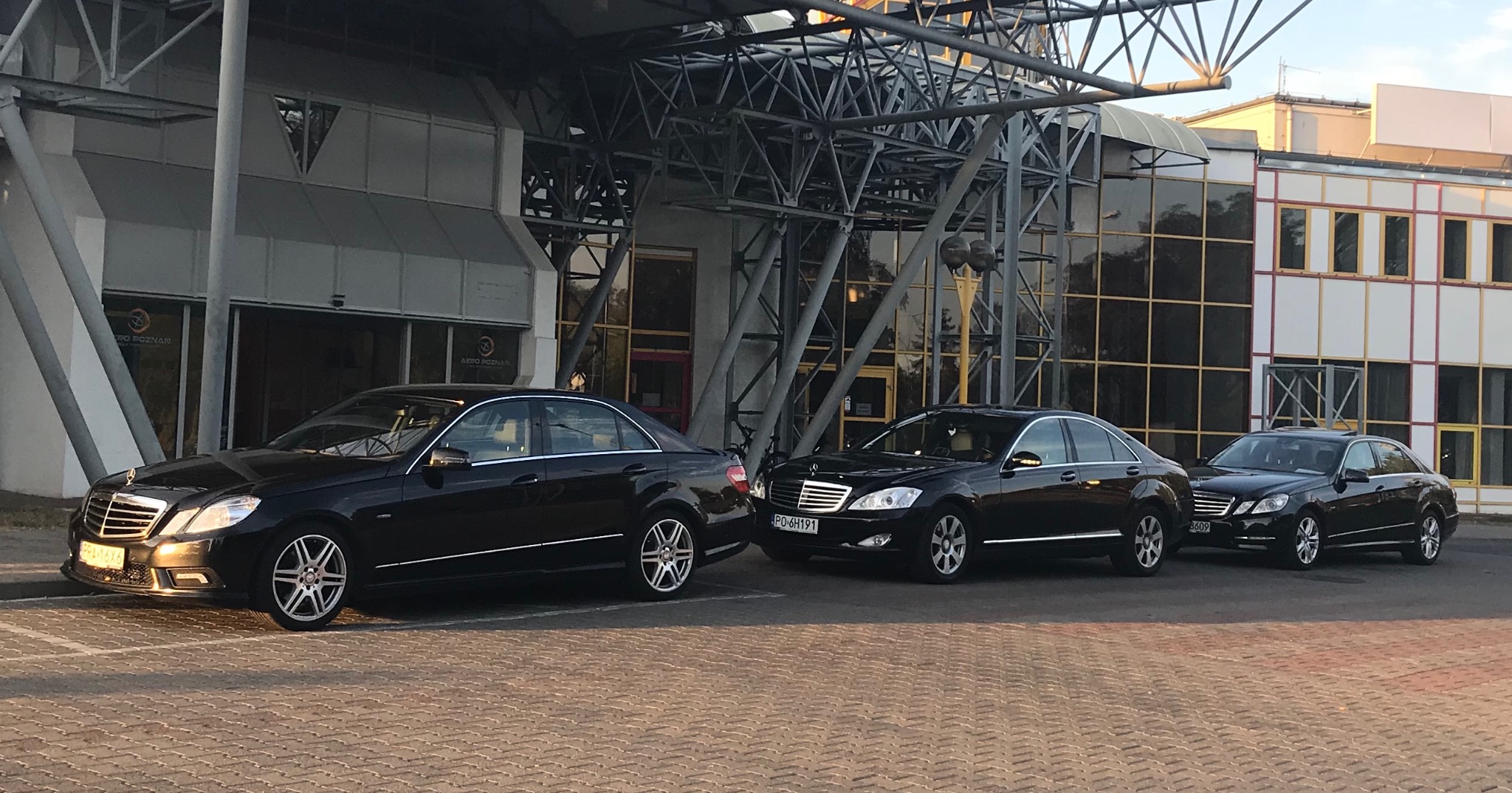 Trzy czarne limuzyny Mercedes-Benz stojące w rzędzie przed nowoczesnym budynkiem z metalową konstrukcją i szklaną fasadą.