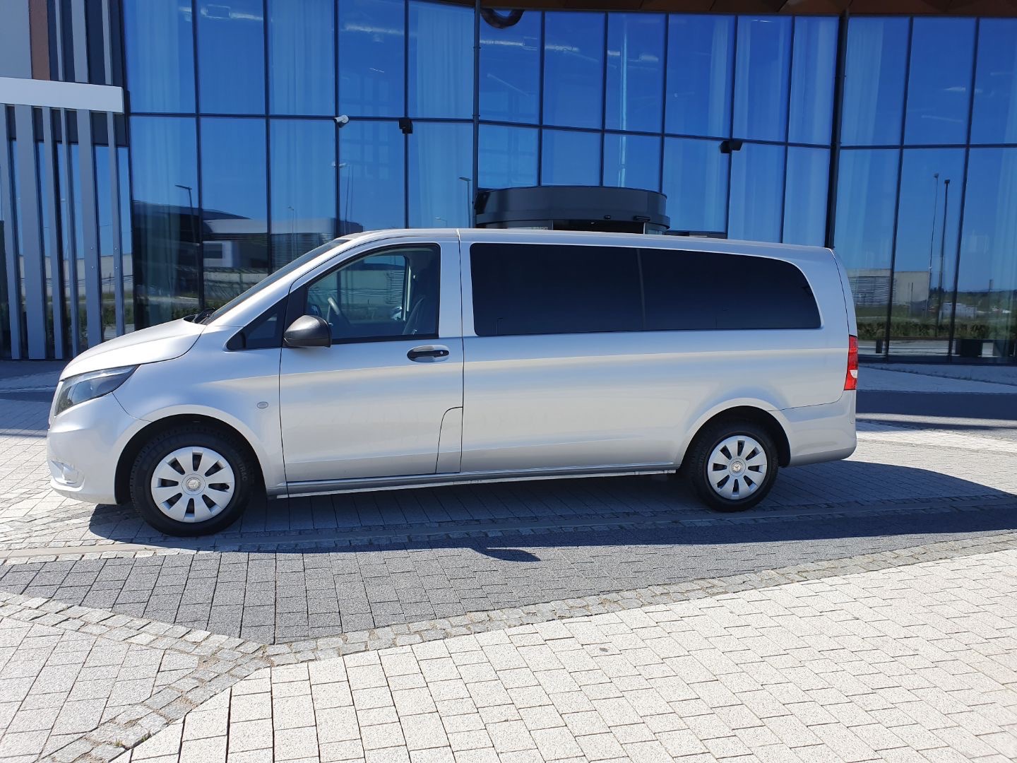 Srebrny Mercedes Vito z bagażnikiem dachowym na tle nowoczesnego budynku biurowego z niebieskimi szybami, zaparkowany na szarej kostce brukowej.