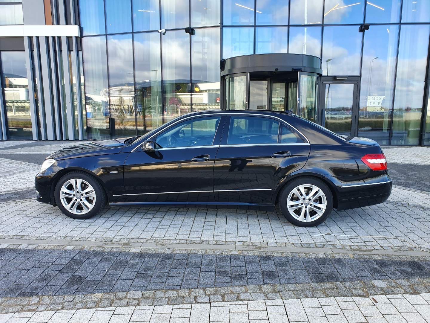 Czarny Mercedes klasy E zaparkowany przed nowoczesnym budynkiem z obrotowymi drzwiami wejściowymi.