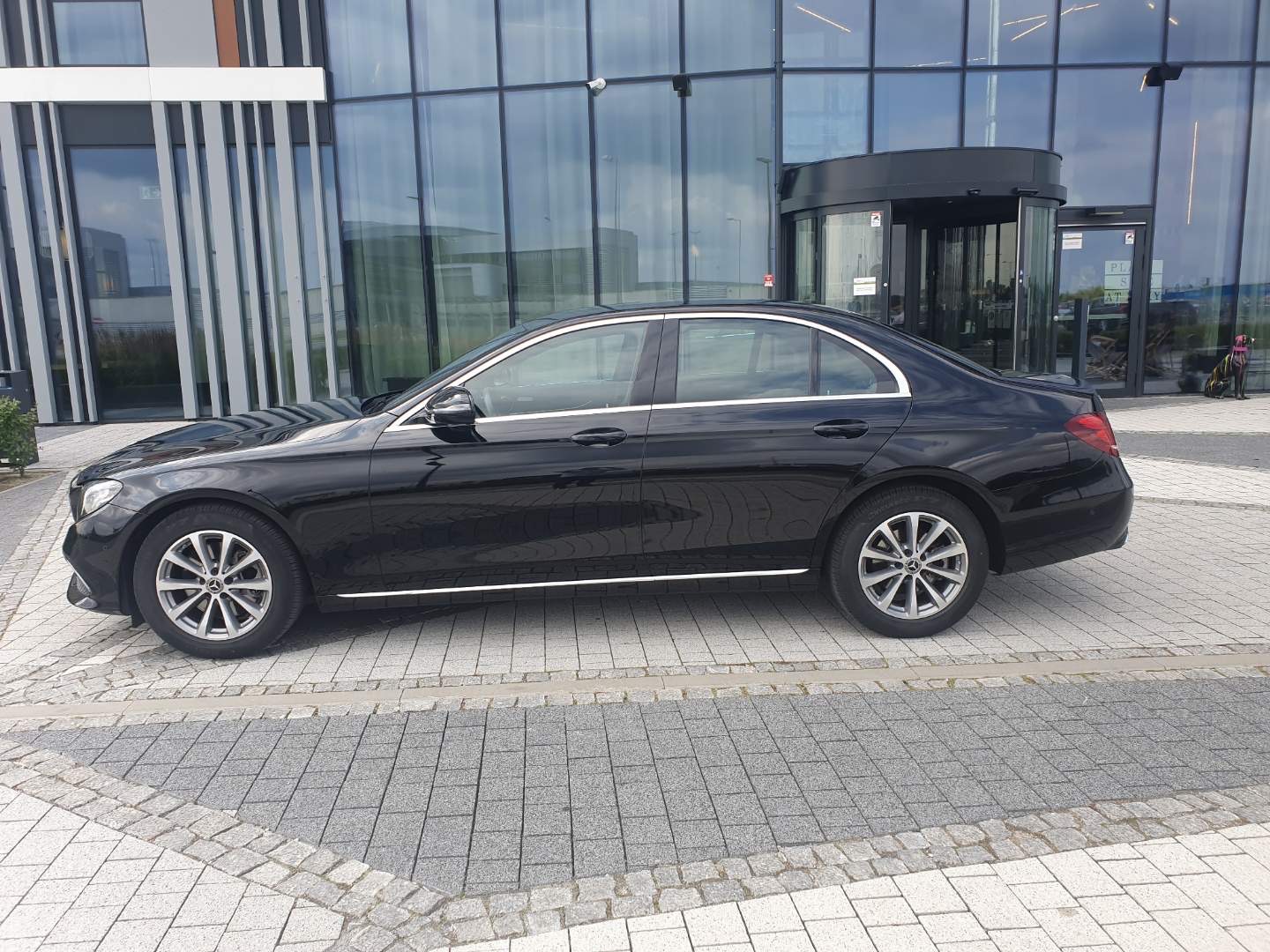 Czarna limuzyna Mercedes zaparkowana przed nowoczesnym budynkiem z obrotowymi drzwiami.