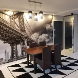 Jadalnia z fototapetą przedstawiającą most w Nowym Jorku, drewnianym stołem i brązowymi krzesłami na czarno-białym dywanie w geometryczne wzory, oświetlona wiszącymi lampami.