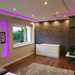 Nowoczesny salon z drewnianą podłogą, ścianą z kamienia dekoracyjnego, białymi meblami i podświetleniem LED w kolorze fioletowym na suficie oraz wnęce ściennej.