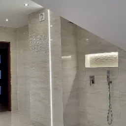 Nowoczesna łazienka na poddaszu z beżowymi płytkami imitującymi marmur, oświetleniem LED w narożniku i wnęce nad prysznicem, oraz widocznym odpływem liniowym w podłodze.