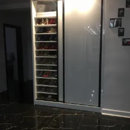 Szafa wnękowa z przesuwnymi, białymi drzwiami i otwartą częścią z półkami na buty, oświetlenie punktowe, podłoga z czarnego marmuru.