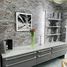 Szara szafka RTV z szufladami i półkami, dekoracje na półkach, kwiaty w wazonie, próbnik kolorów na blacie, ściana z efektem starej cegły, czarno-białe zdjęcie w ramce.