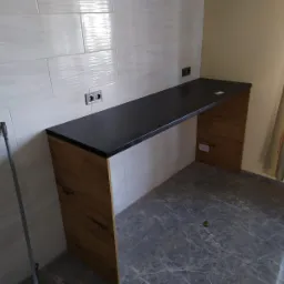 Wysoki blat kuchenny z czarnym blatem i drewnopodobną obudową, zamontowany przy ścianie z białych płytek, widoczne gniazdka elektryczne, podłoga z szarego marmuru.