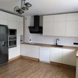 Jasna kuchnia z białymi lakierowanymi frontami, drewnianymi blatami i obudową lodówki, czarnym okapem i lampą sufitową w stylu retro, płytki imitujące cegłę na ścianie.