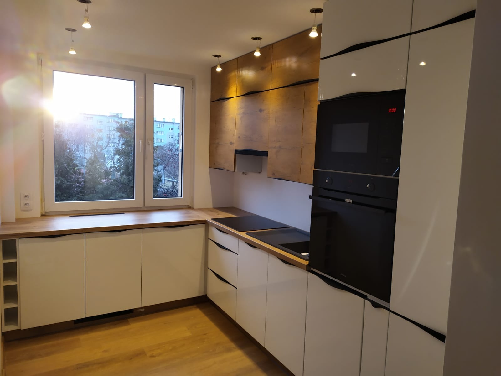 Nowoczesna kuchnia z białymi szafkami dolnymi, drewnianym blatem, i górnymi szafkami w kolorze drewna z czarnymi akcentami, oświetlona światłem słonecznym wpadającym przez okno.