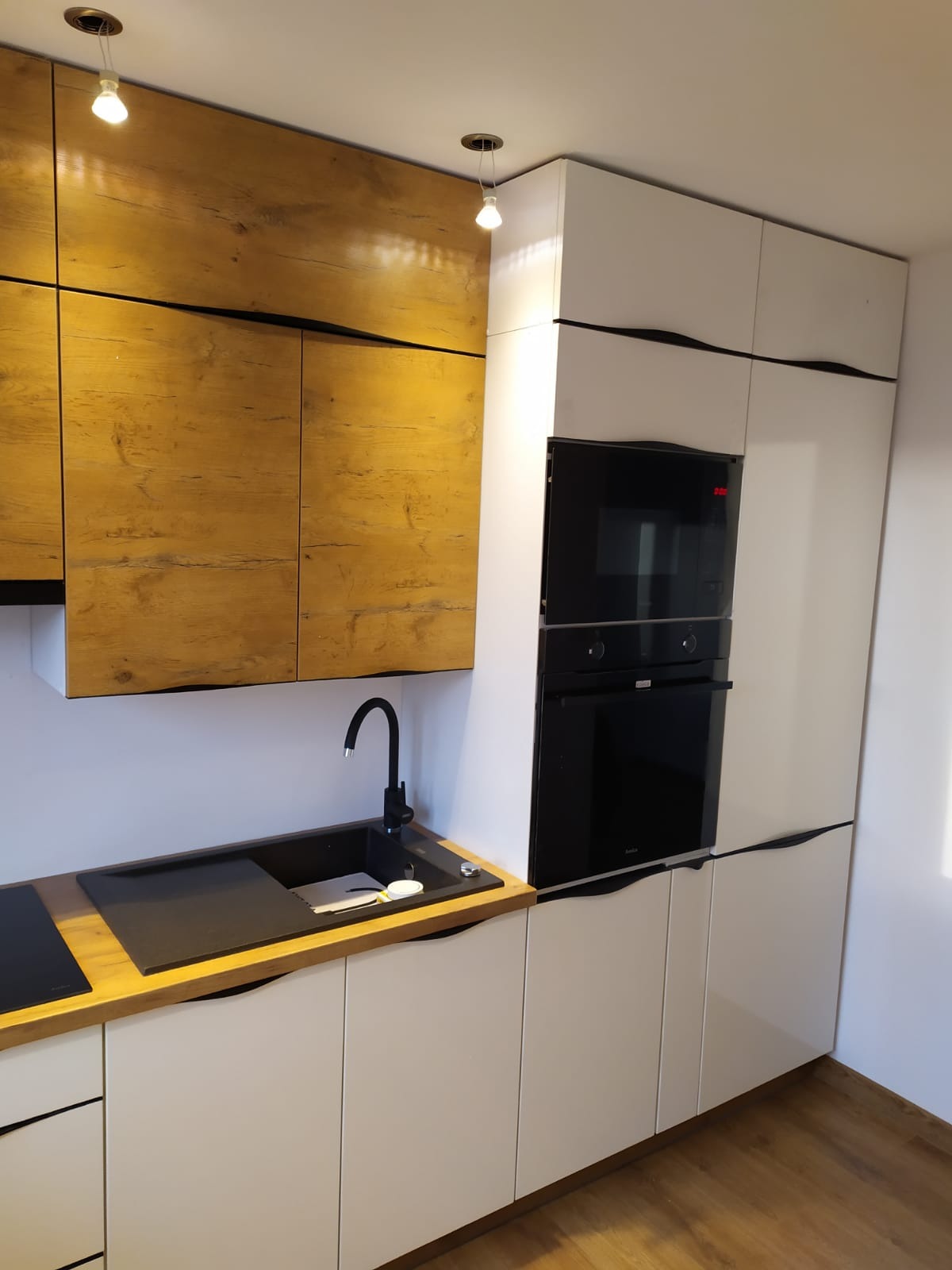 Nowoczesna kuchnia: białe szafki z frezowanymi uchwytami, drewniane blaty, czarny zlew z baterią i zabudowane piekarniki.