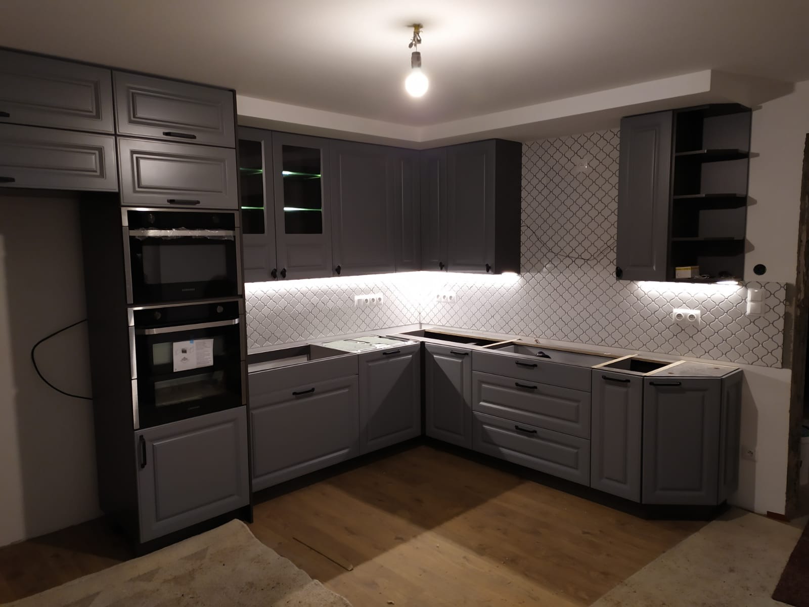 Szara kuchnia w trakcie montażu, widoczne szafki górne z przeszkleniami, zabudowa piekarnika i płyty grzewczej, ozdobna mozaika na ścianie, brak blatów.