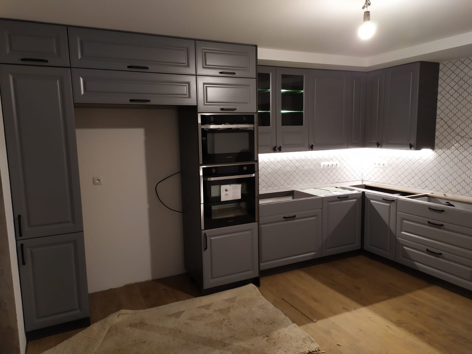 Szara zabudowa kuchenna z piekarnikiem i mikrofalówką w trakcie montażu, widoczne niezabezpieczone blaty i brakujące elementy wykończeniowe, podświetlenie LED nad blatem.