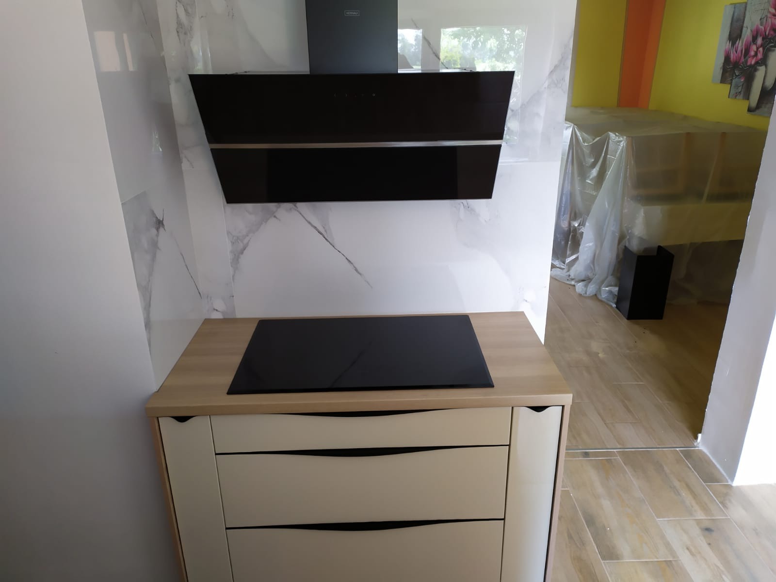 Fragment kuchni z płytą indukcyjną w blacie i czarnym okapem, marmurowe płytki na ścianie, w tle meble zabezpieczone folią malarską.