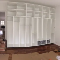 Wielka biała konstrukcja regałowa z licznymi otwartymi półkami i szufladami w trakcie montażu, widoczne metalowe prowadnice szuflad, na podłodze rozłożone kartony zabezpieczające.