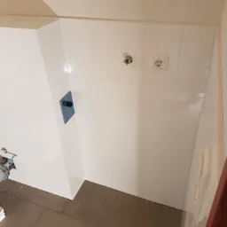 Białe, błyszczące płytki ścienne w łazience, z widocznym gniazdkiem elektrycznym i wystającym przyłączem wodnym, brązowe płytki podłogowe, częściowo widoczne narzędzia glazurnicze.