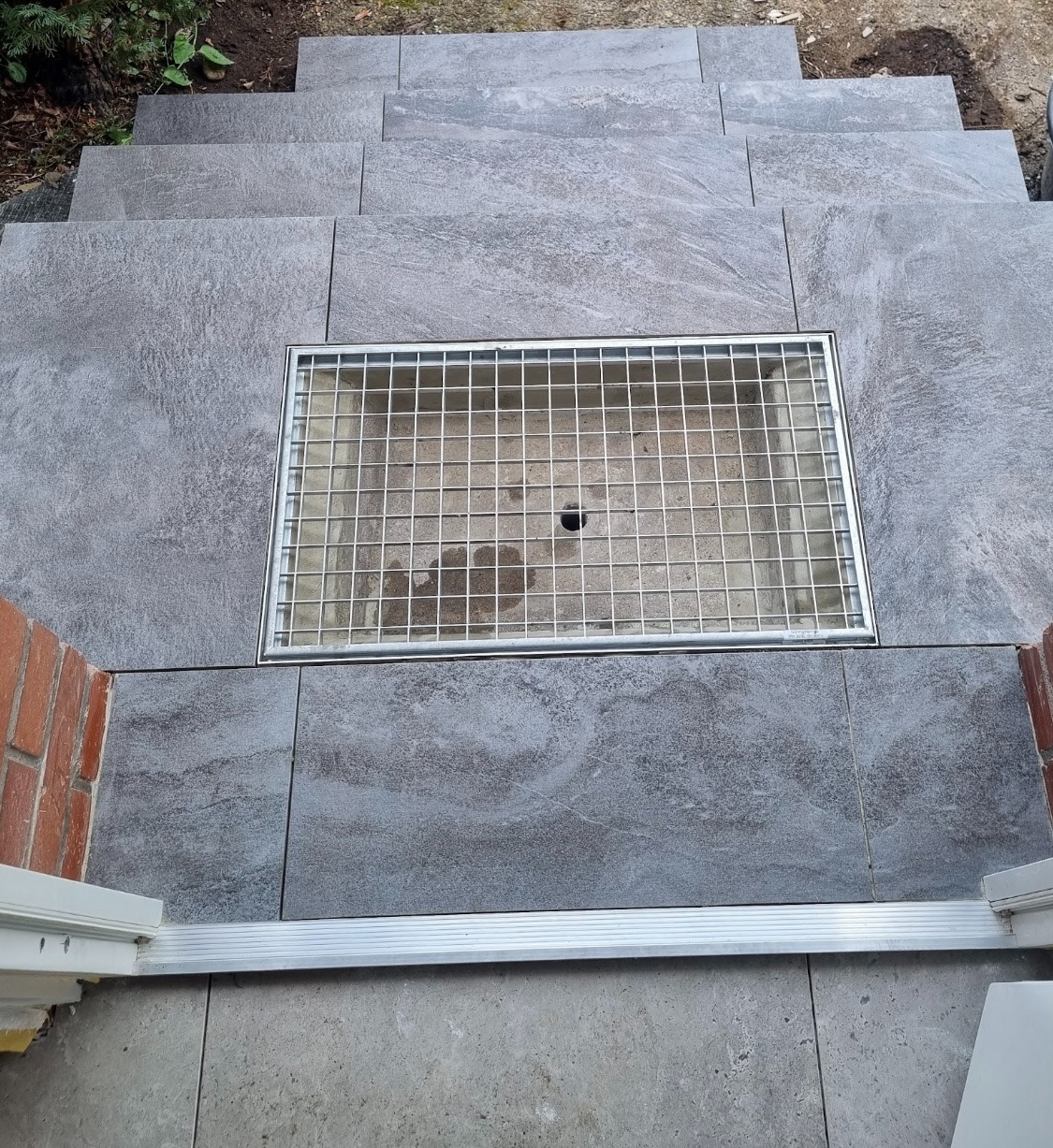 Schody zewnętrzne wyłożone szarymi płytkami imitującymi kamień, z wbudowaną metalową kratką odpływową, widok z góry z perspektywy wejścia.