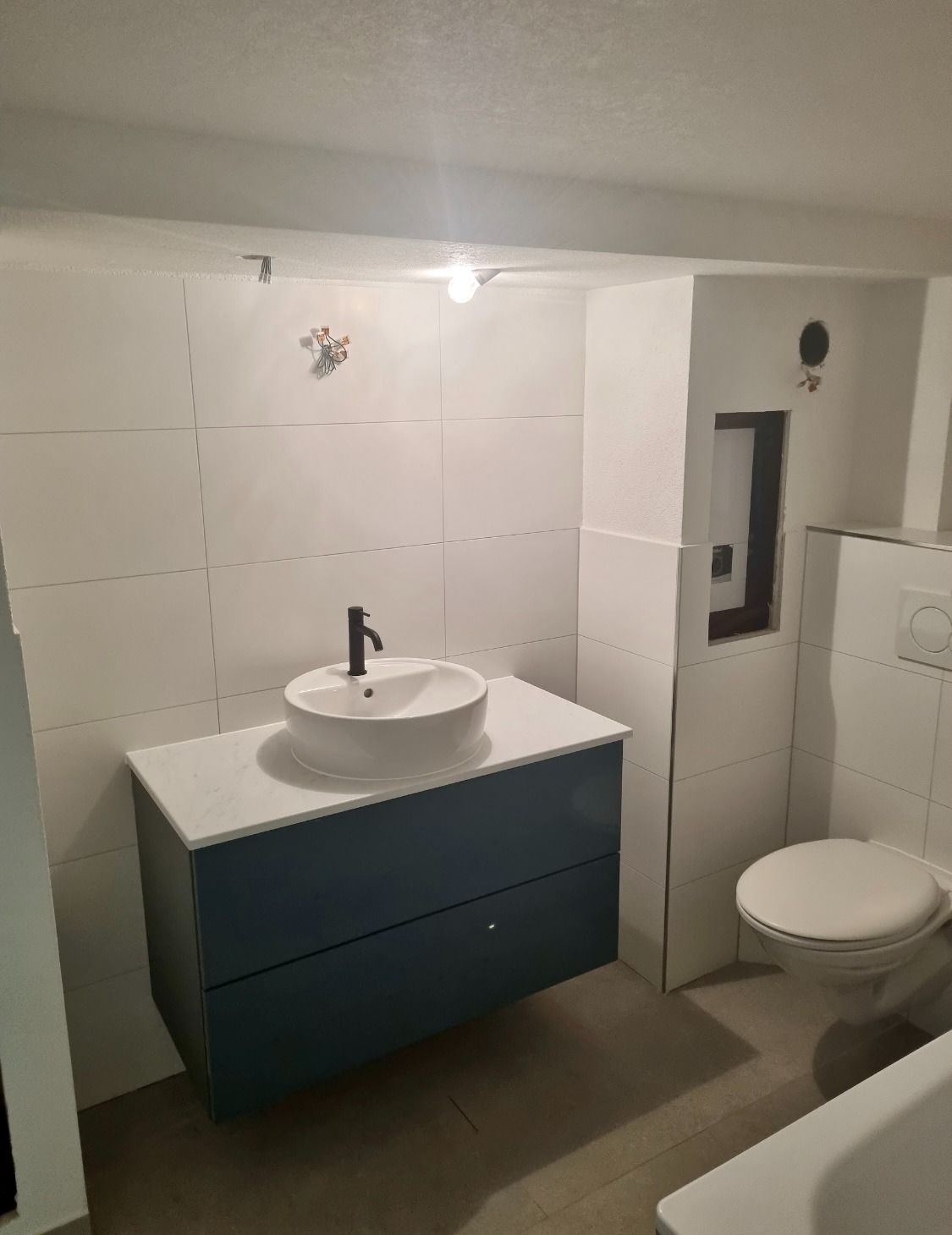 Nowoczesna łazienka z umywalką nablatową na szafce w kolorze granatowym, białe płytki na ścianach i widoczne niedokończone elementy instalacji elektrycznej.