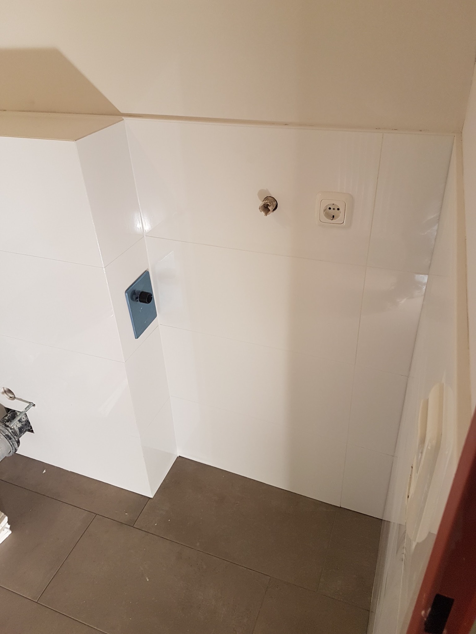 Białe, błyszczące płytki ścienne w łazience, z widocznym gniazdkiem elektrycznym i wystającym przyłączem wodnym, brązowe płytki podłogowe, częściowo widoczne narzędzia glazurnicze.