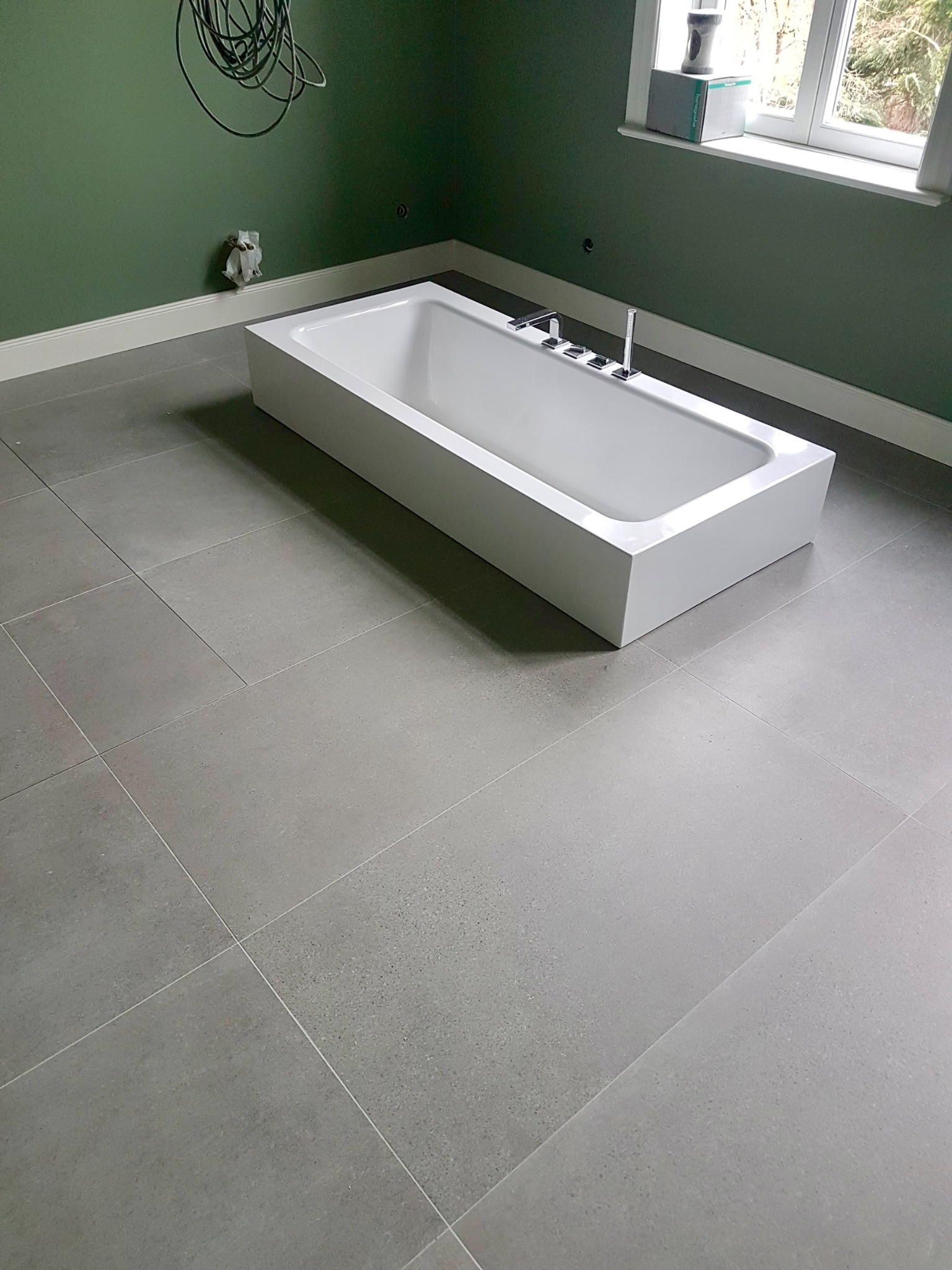 Minimalistyczna, biała wanna wbudowana w szare płytki podłogowe, zielona ściana z widocznymi przewodami i pudełko na parapecie okiennym.