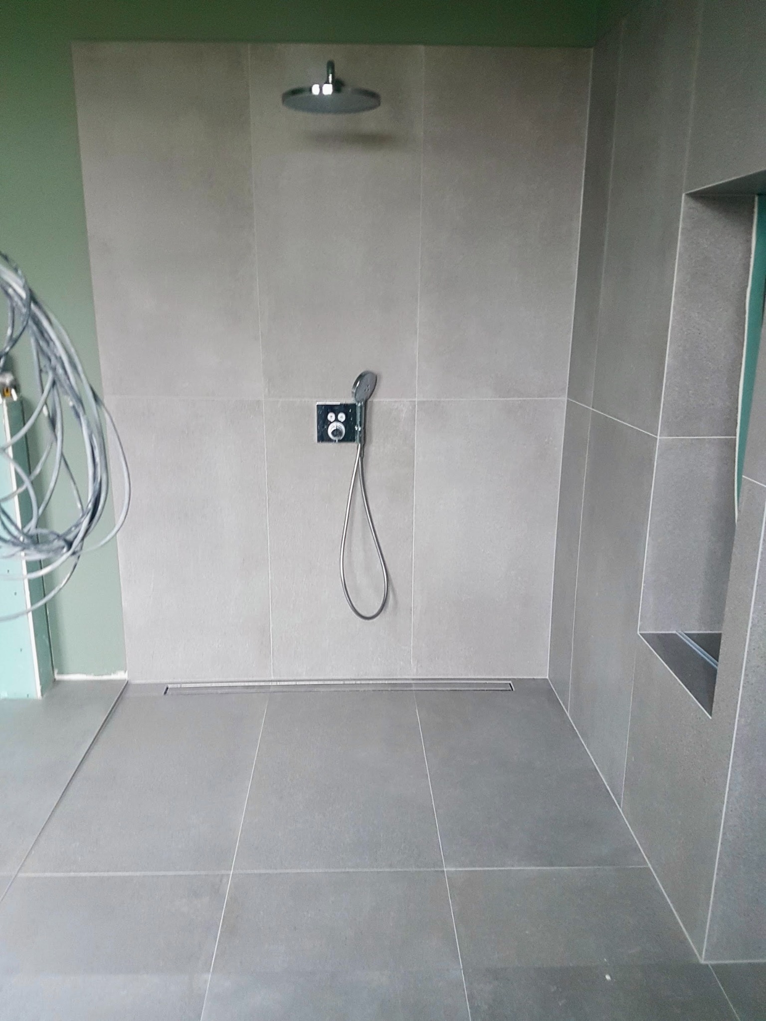 Szara łazienka w trakcie wykończenia, widoczne płytki na ścianach i podłodze, deszczownica, panel prysznicowy z słuchawką, odpływ liniowy w podłodze, ściana z niedokończonym wykończeniem...