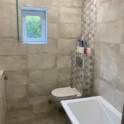 Wykończona łazienka z beżowymi płytkami, wiszącą toaletą, białą wanną i oknem z widokiem na zieleń.