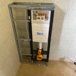 Stelaż podtynkowy WC marki Roca zamontowany w metalowej konstrukcji, widoczne żółte elementy instalacji kanalizacyjnej, płytki ceramiczne na ścianach, przygotowanie do wylewki na podłodze...
