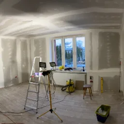 Pokój w trakcie prac wykończeniowych, ściany i sufit pokryte płytami gipsowo-kartonowymi, widoczna drabina, lampy budowlane, odkurzacz i narzędzia.