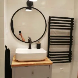 Nowoczesna łazienka z białą umywalką nablatową na szarej szafce z drewnianym blatem, okrągłe lustro w czarnej ramie odbija rękę robiącą zdjęcie, czarny grzejnik drabinkowy na białej ścianie...