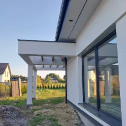 Nowoczesny dom jednorodzinny z białą elewacją, dużym oknem przesuwnym i zadaszeniem tarasu z drewnianymi elementami, widok z zewnątrz na ogród i sąsiednie budynki.
