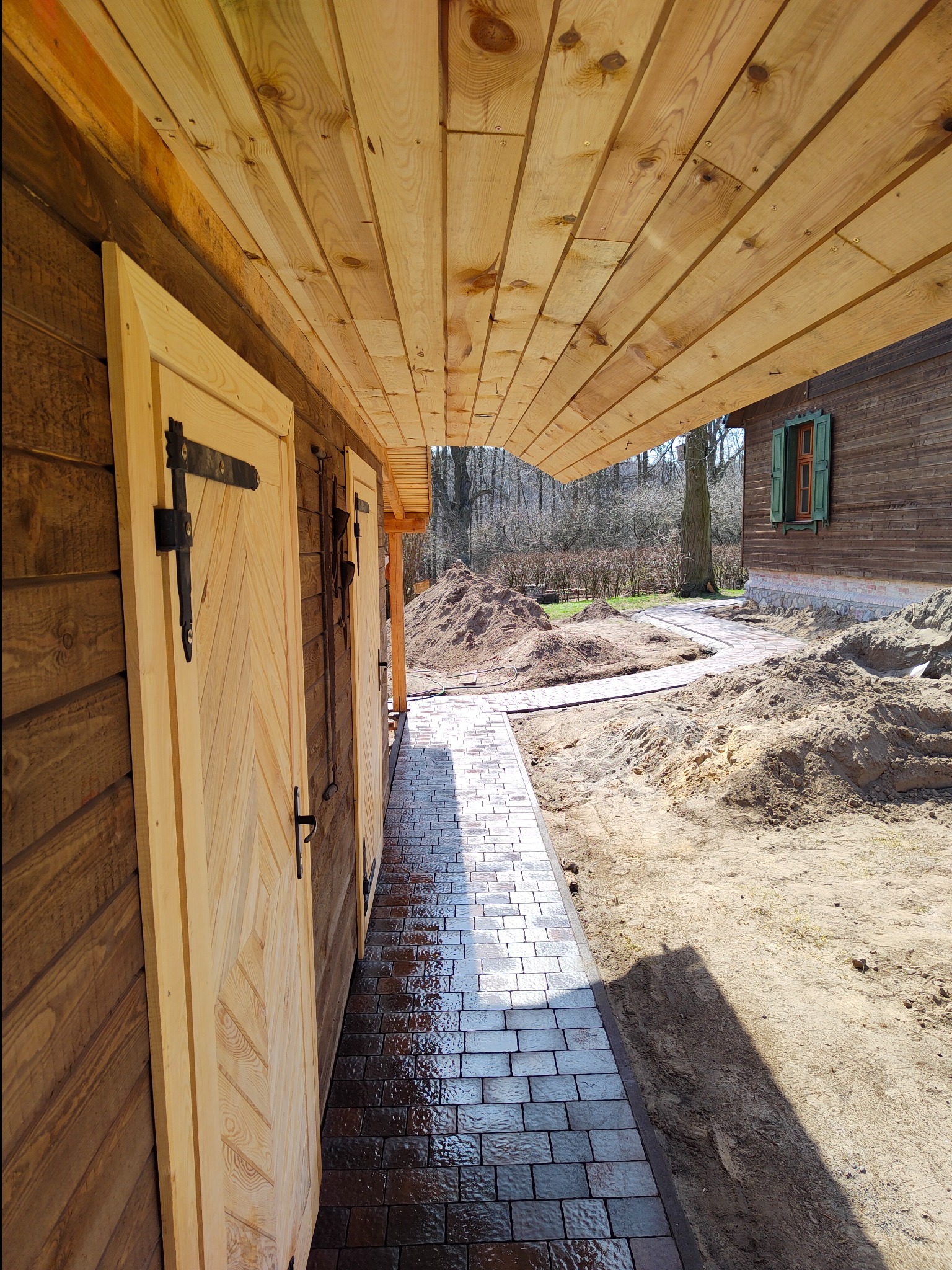Świeżo ułożona, mokra kostka brukowa prowadząca wzdłuż drewnianego budynku z jasnymi, ozdobnymi drzwiami i drewnianym podbitkiem dachu, w tle hałda ziemi i drugi budynek z zielonymi okiennicami.
