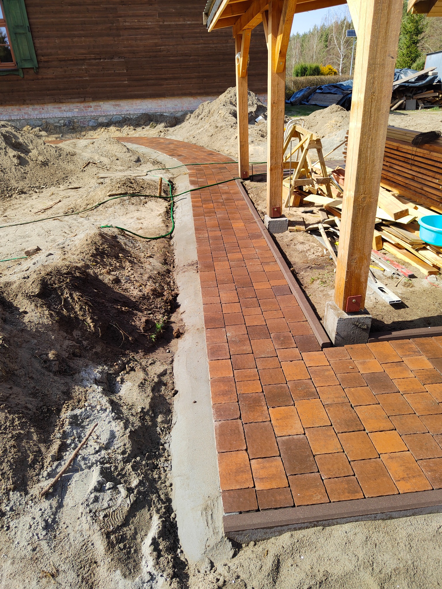 Ścieżka z pomarańczowo-brązowej kostki brukowej, wijąca się obok drewnianej konstrukcji z widocznymi deskami i narzędziami, otoczona piaskiem i ziemią, w trakcie budowy.