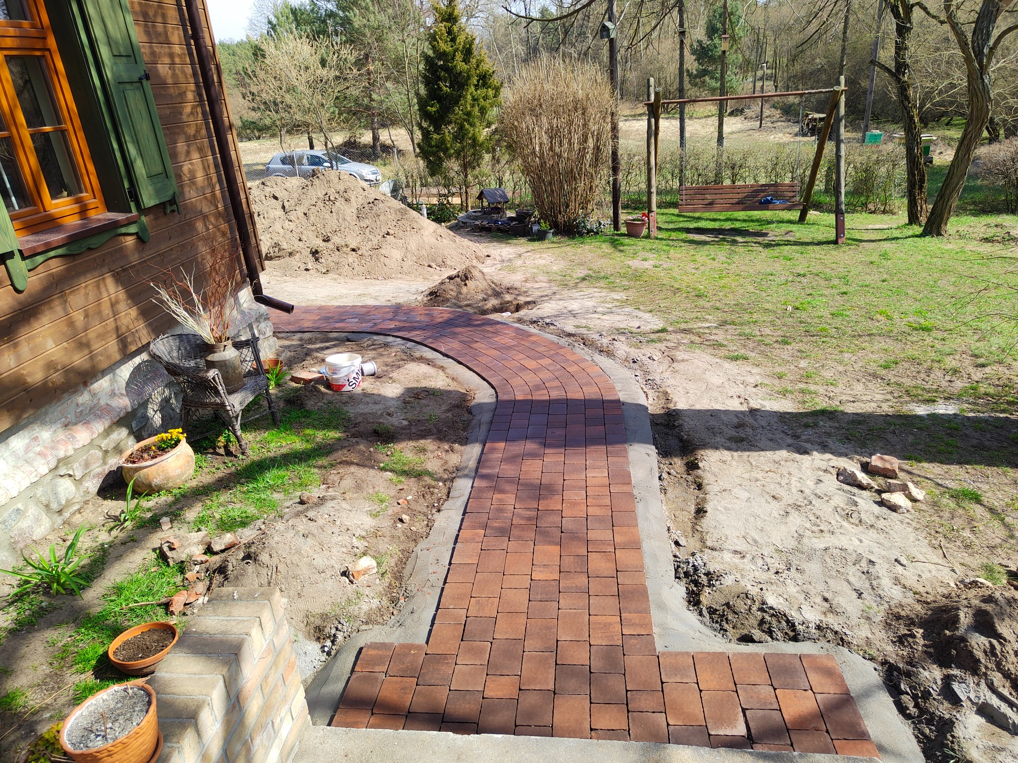 Czerwona kostka brukowa ułożona w ścieżkę prowadzącą od schodów domu w kierunku ogrodu z huśtawką, widoczny nasyp ziemi i elementy krajobrazu w tle.