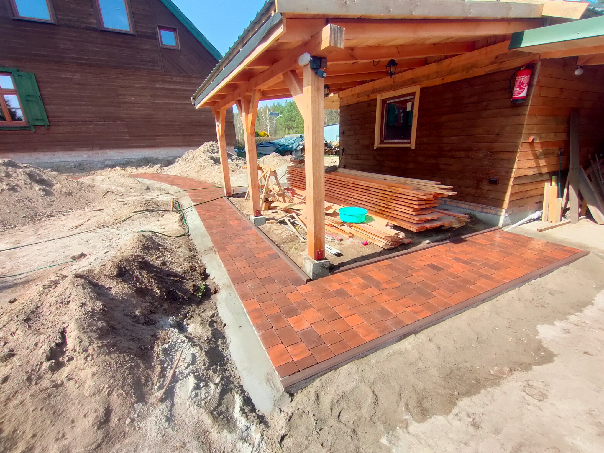 Czerwona kostka brukowa ułożona wzdłuż ścieżki i tarasu przy drewnianym domu z zadaszeniem, widoczne przygotowane deski i ziemia do wyrównania terenu.