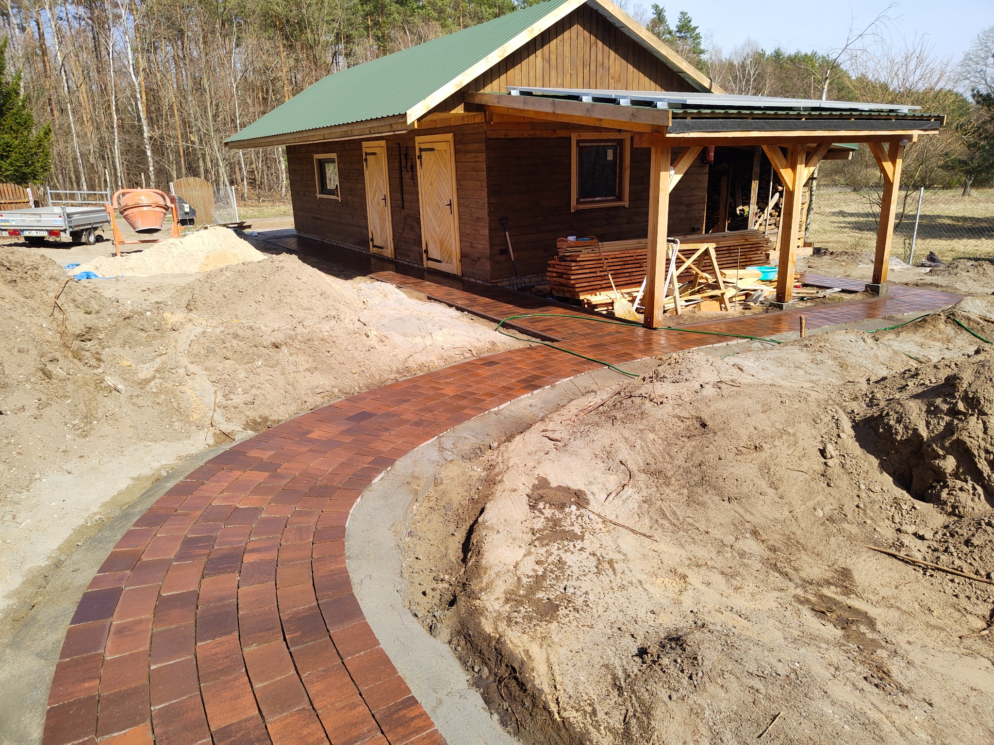 Kręta ścieżka z czerwonej kostki brukowej prowadząca od drewnianego domku z zielonym dachem, widoczny wykop pod ścieżkę z obrzeżami, w tle betoniarka i przyczepa.