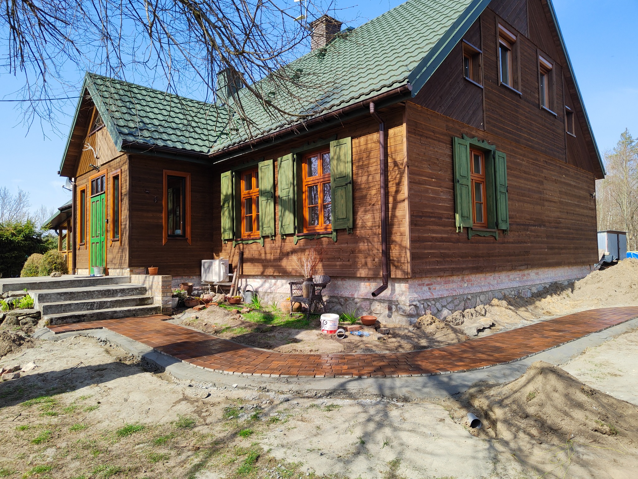 Ułożona ścieżka z kostki brukowej prowadząca do drewnianego domu z zielonym dachem, widoczny fragment ogrodu w trakcie prac wykończeniowych.