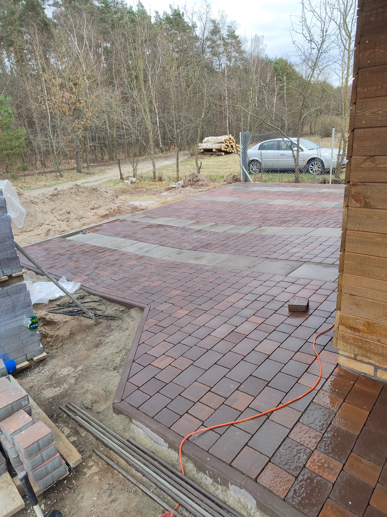 Ułożona nawierzchnia z kostki brukowej w odcieniach brązu i szarości, z widocznymi elementami w trakcie prac wykończeniowych, takimi jak stosy kostki, rury i przewód elektryczny, na tle naturalnego...
