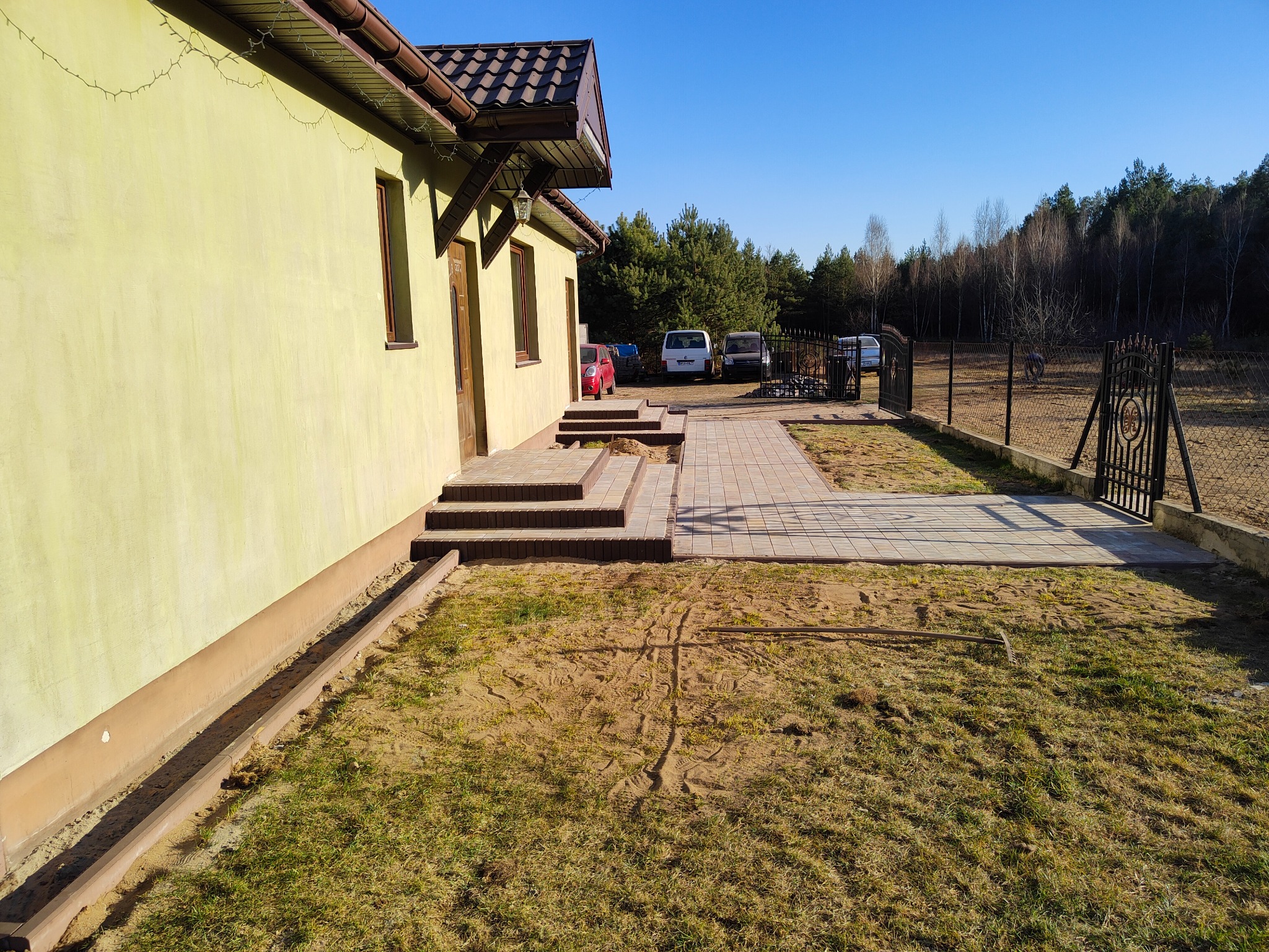 Ułożona kostka brukowa prowadząca od domu z schodami do bramy wjazdowej, widoczne ślady prac ziemnych na trawniku, w tle zaparkowane samochody i las.
