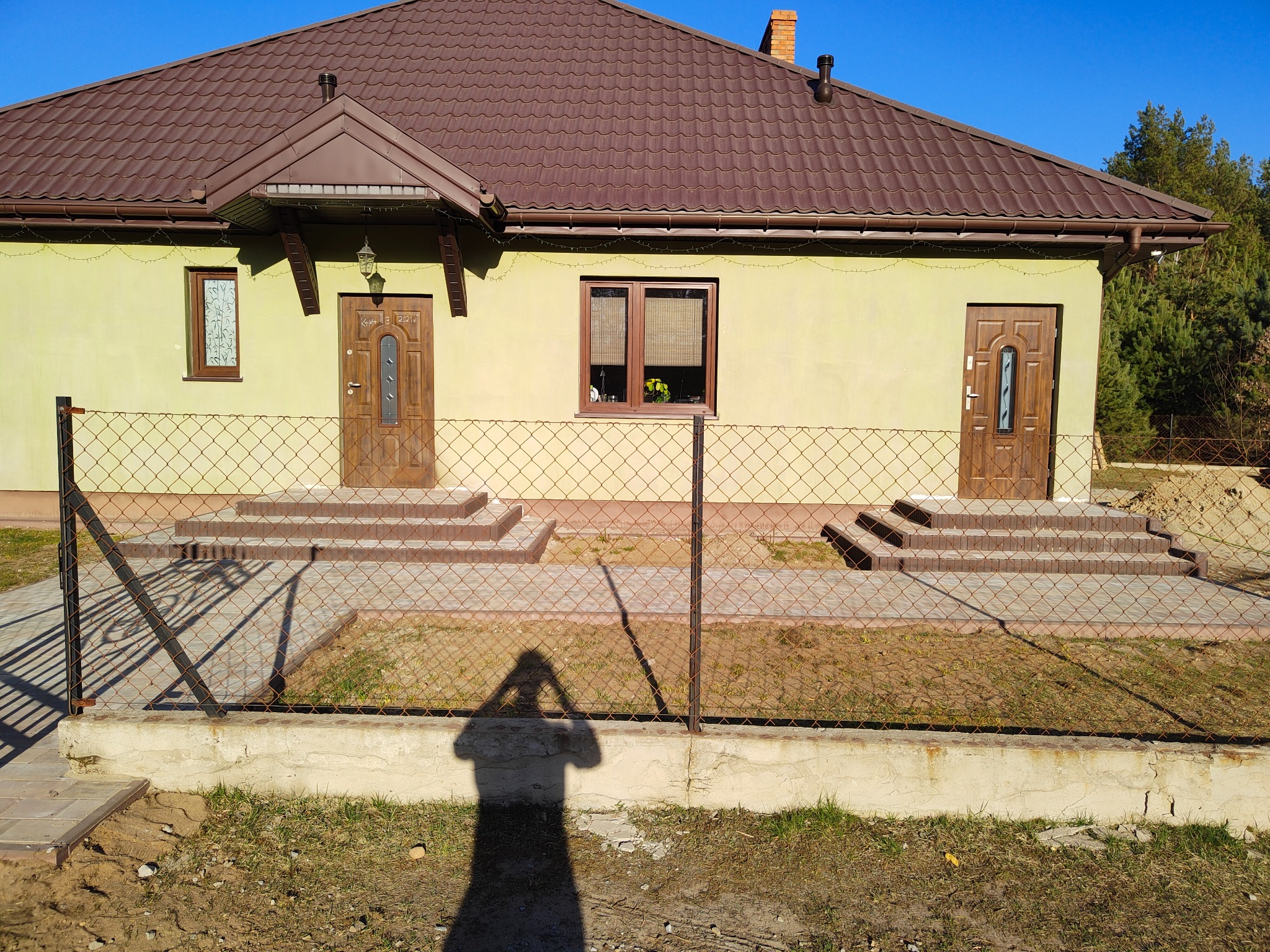 Nowo wybudowany dom jednorodzinny z brązowym dachem i dwoma wejściami, widoczny przez siatkę ogrodzeniową, cień osoby fotografującej na pierwszym planie.