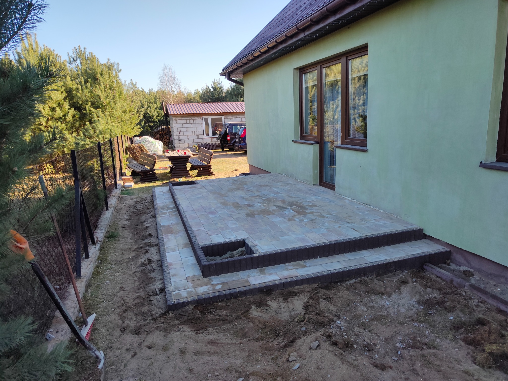 Niedokończony taras z kostki brukowej z dwoma stopniami, obrzeża z ciemniejszej kostki, widoczne narzędzia ogrodnicze i świeżo rozkopana ziemia wokół, dom z zieloną elewacją w tle.