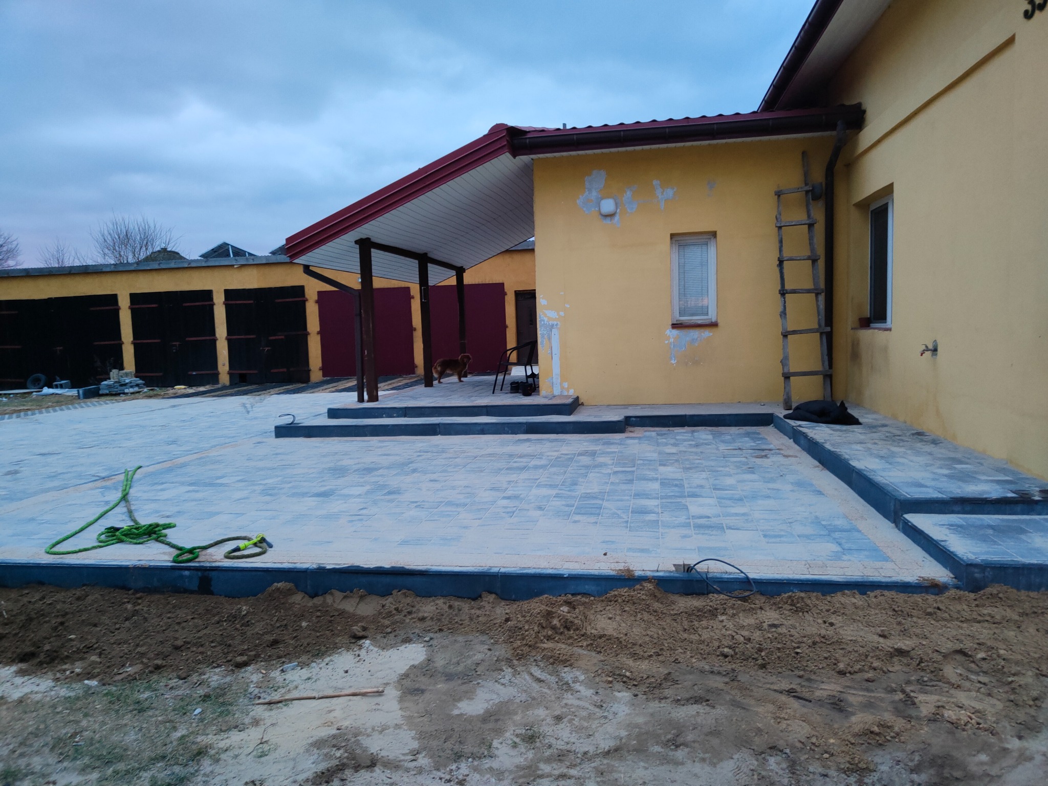 Niedawno ułożona szara kostka brukowa przed żółtym domem z drewnianą drabiną opartą o ścianę, widoczna lina na kostce brukowej i ziemia wzdłuż krawędzi.