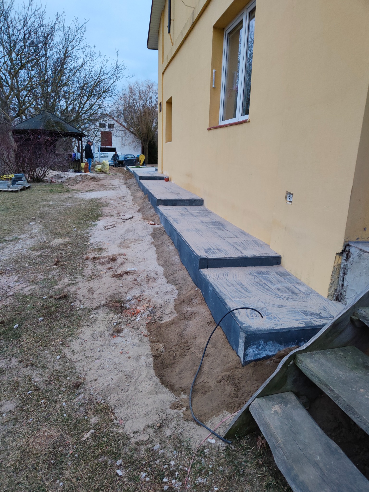 Układanie stopni z kostki brukowej przy żółtej ścianie budynku, widoczne niedociągnięcia w obróbce i resztki piasku na powierzchni, w tle altana i osoby.