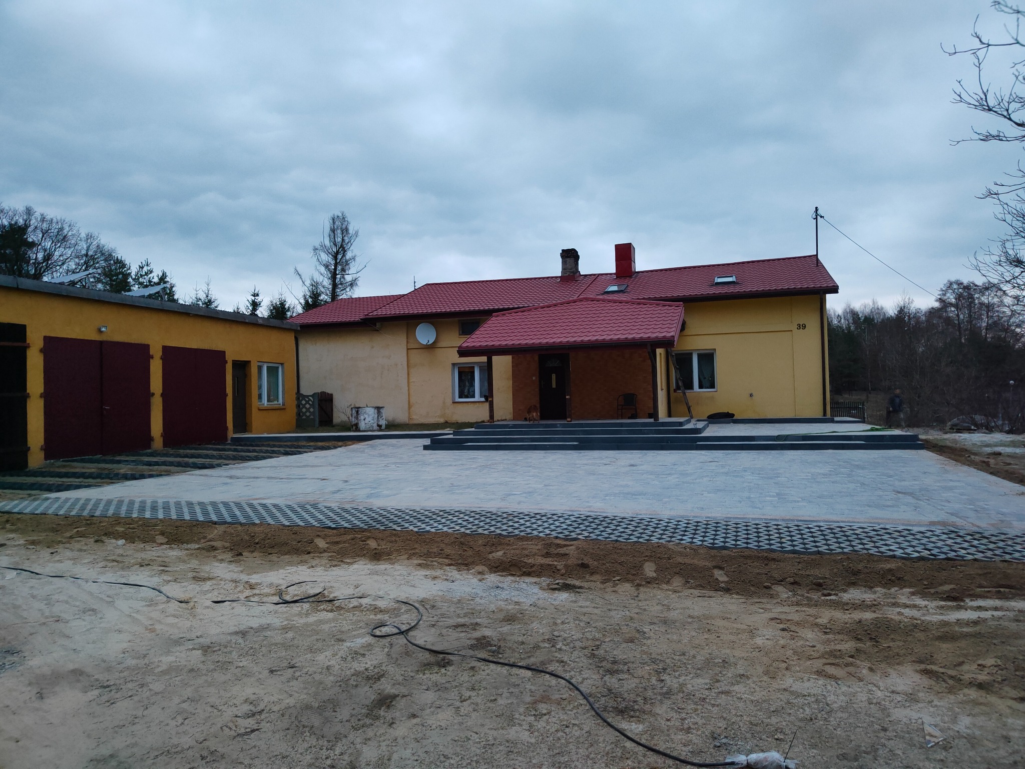 Nowo ułożony, szary podjazd z kostki brukowej ażurowej i pełnej przed żółtym budynkiem z czerwoną dachówką w pochmurny dzień. Widoczne ślady piasku i ziemi po pracach wykończeniowych.