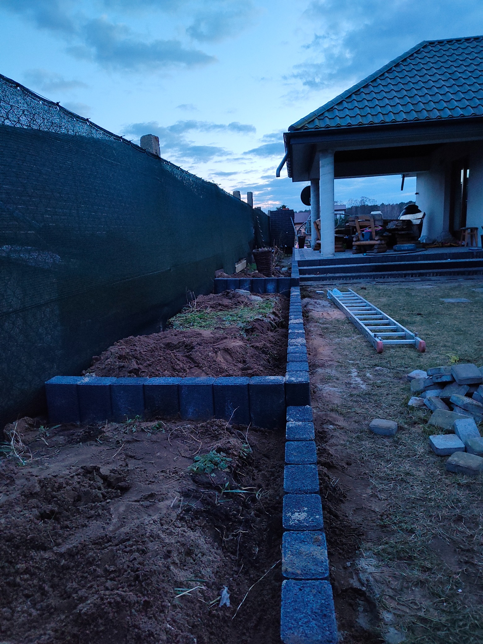 Wykop pod obrzeża rabaty z ułożoną częścią krawężnika z kostki betonowej, ziemia wysypana wzdłuż ogrodzenia z siatki maskującej, w tle drabina oparta o budynek gospodarczy.