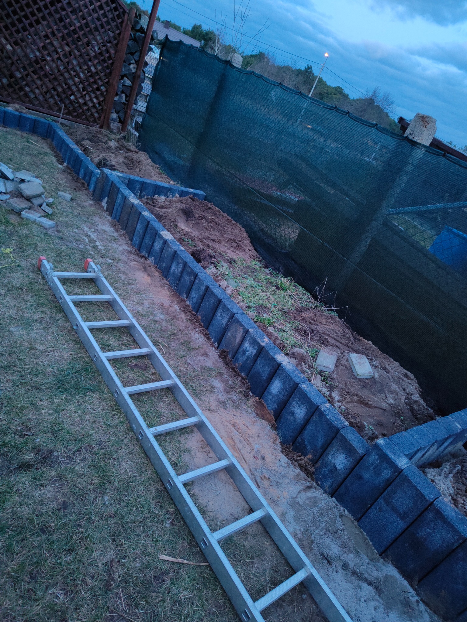 Wykop ziemny z częściowo ułożonym obrzeżem z betonowych kostek wzdłuż ogrodzenia, na trawie leży aluminiowa drabina, widoczne prace przygotowawcze.