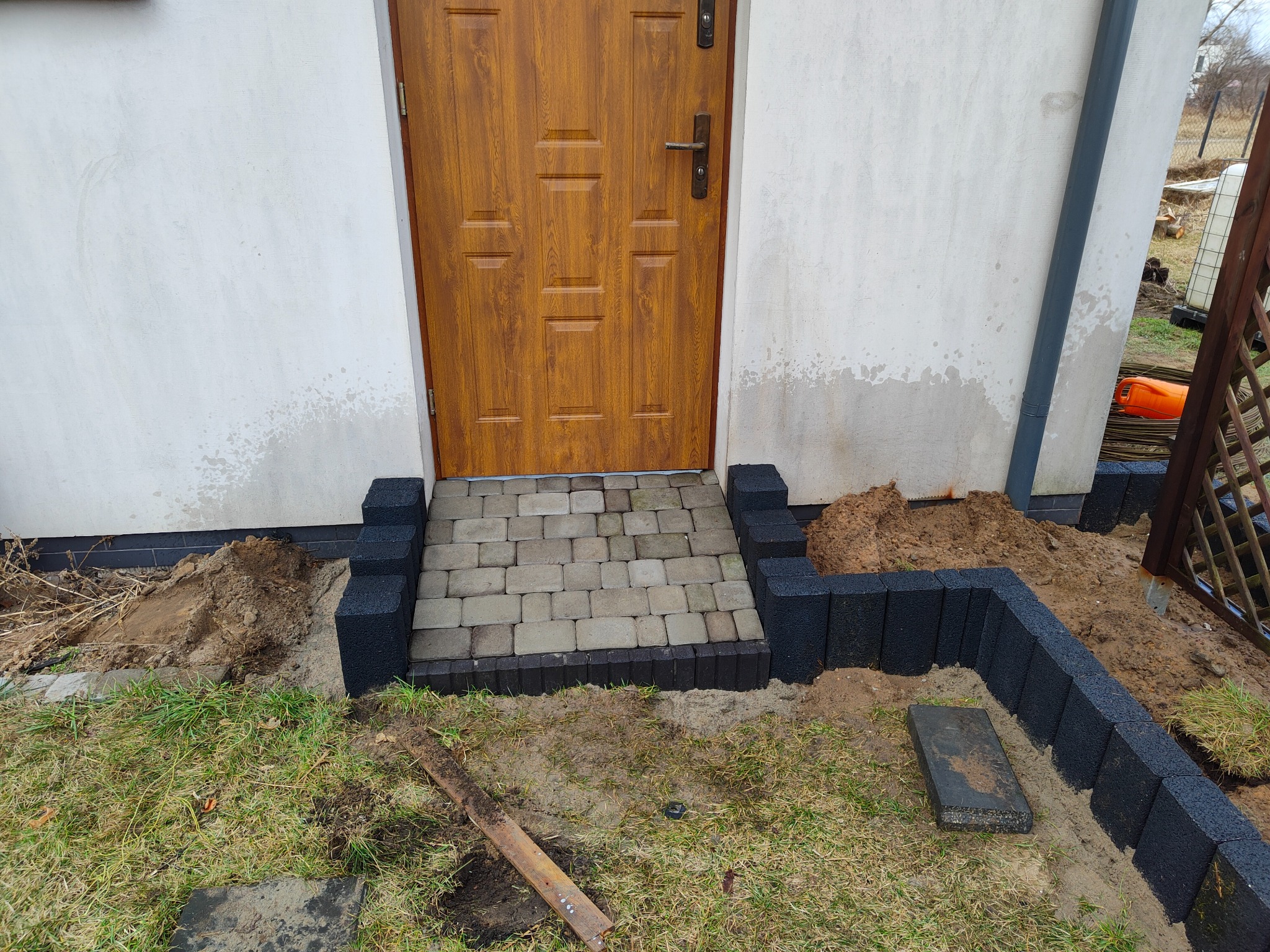 Wejście do domu z drewnianymi drzwiami, wyłożone jasną kostką brukową obramowaną czarnymi bloczkami, widoczne ślady prac ziemnych i trawa.