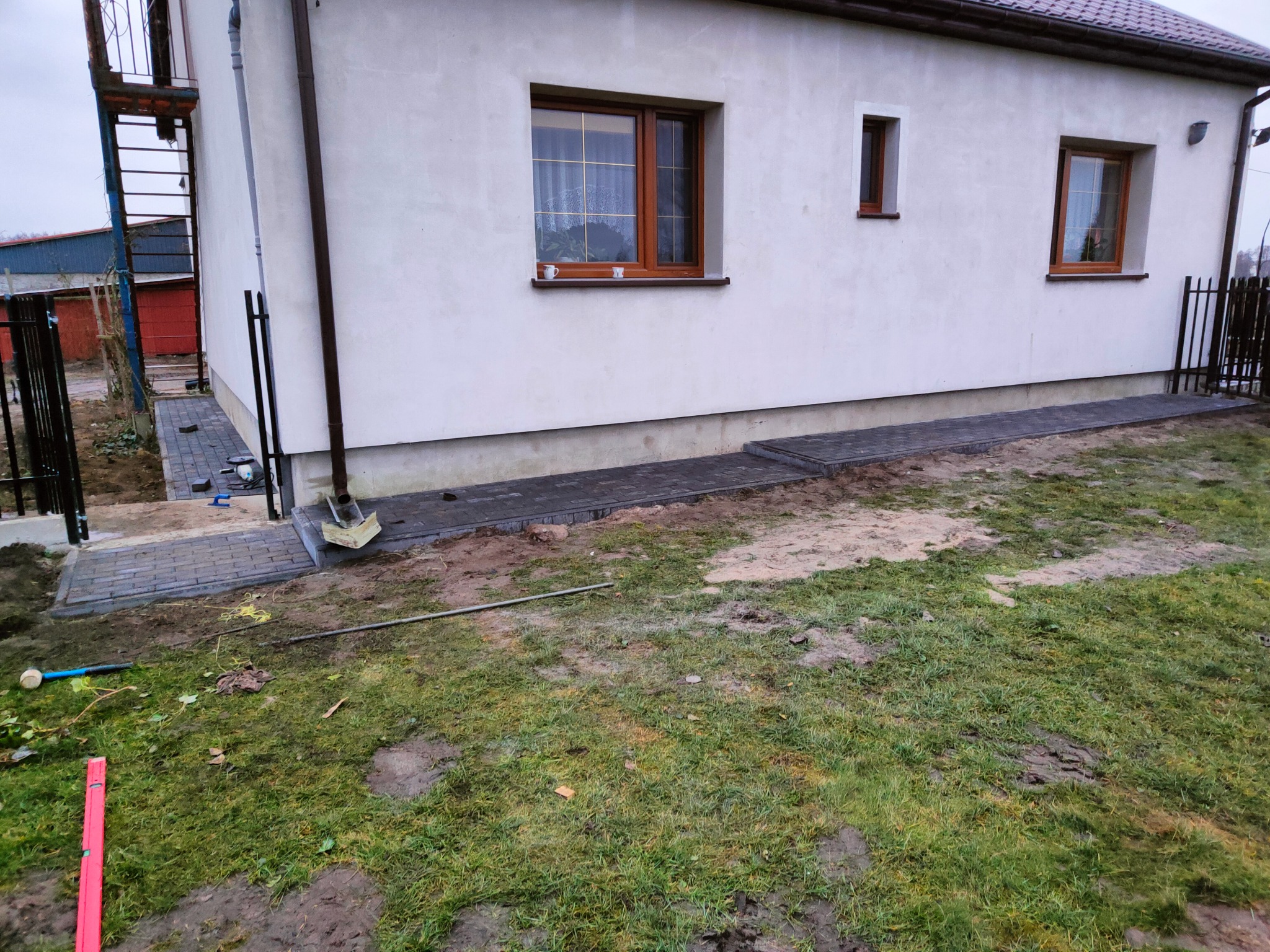 Ułożona ścieżka z szarej kostki brukowej prowadząca od schodów do budynku z białymi ścianami i brązowymi oknami, widoczne narzędzia brukarza i trawnik z piaskiem.