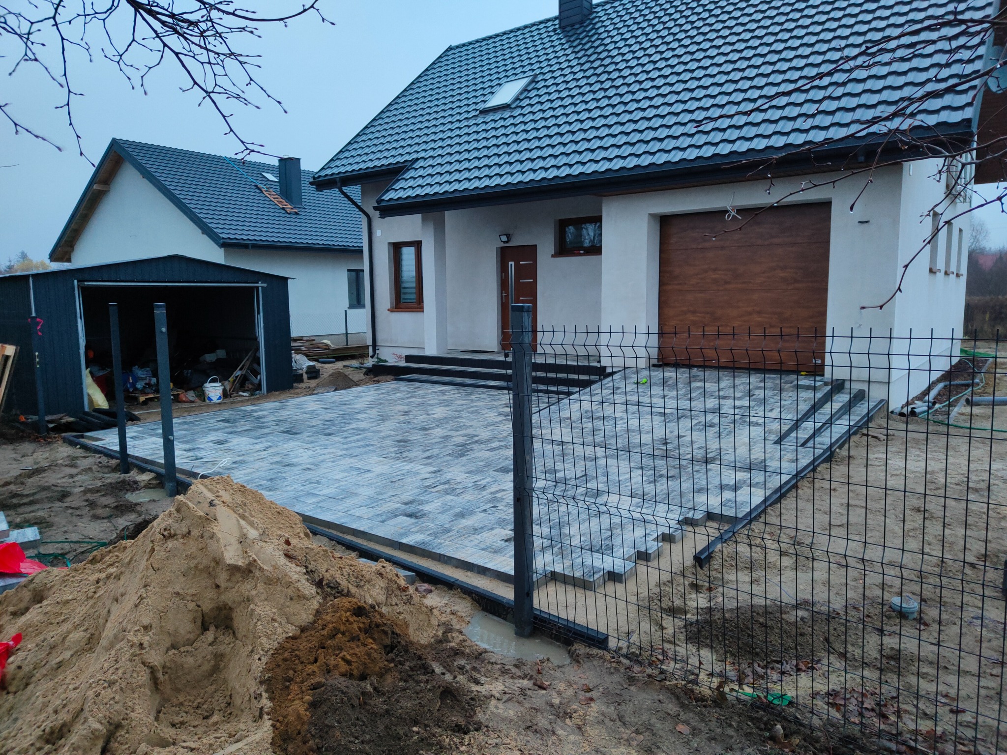 Układanie szarej kostki brukowej na podjeździe przed nowoczesnym domem z brązową bramą garażową, widoczny fragment ogrodzenia i sterta piasku.