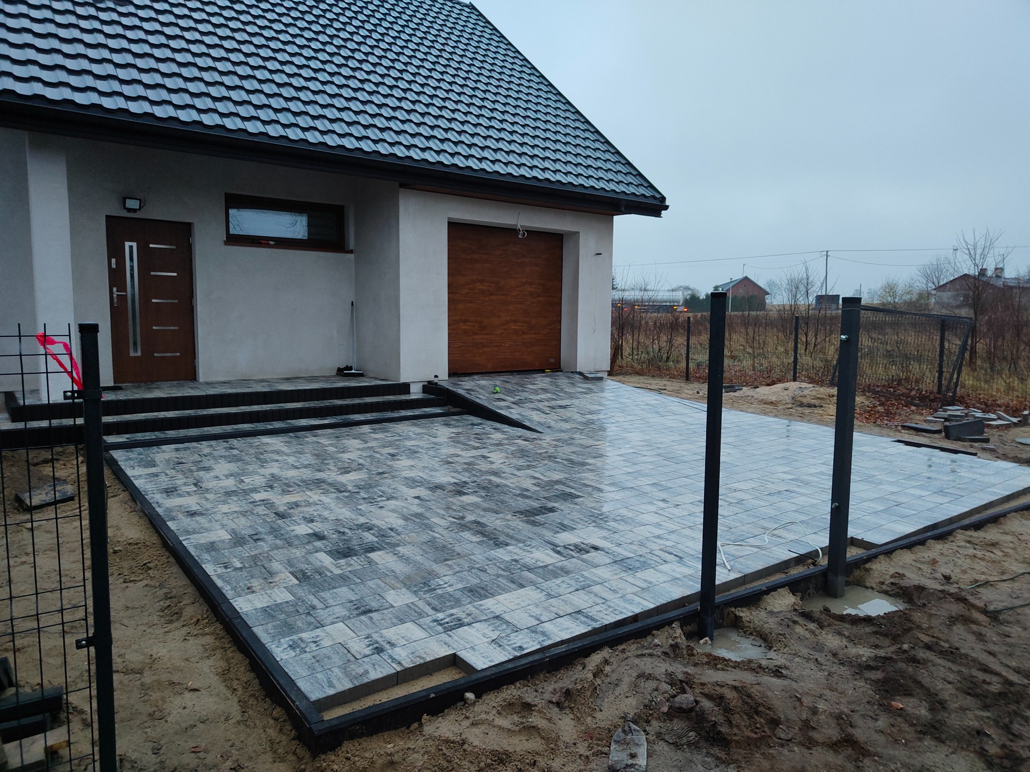 Świeżo ułożona, mokra nawierzchnia z szaro-białej kostki brukowej przed nowoczesnym domem z grafitowym dachem, widoczne obrzeża i przygotowane otwory na słupki ogrodzeniowe.